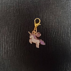 May include: A gold-tone charm featuring a white unicorn with a purple mane and tail. The charm has a small purple gem and a gold-tone lobster clasp.