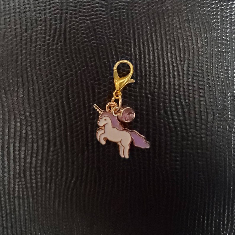 May include: A gold-tone charm featuring a white unicorn with a purple mane and tail. The charm has a small purple gem and a gold-tone lobster clasp.