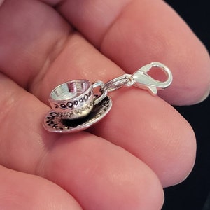 May include: A silver-colored charm featuring a miniature teacup and saucer. The teacup has a decorative pattern around the rim and a small handle. The charm is attached to a lobster clasp for easy attachment to a bracelet or necklace.