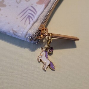 May include: A white zippered pouch with a floral pattern and a gold zipper pull. The zipper pull has a small unicorn charm with a purple mane and a pink gem.