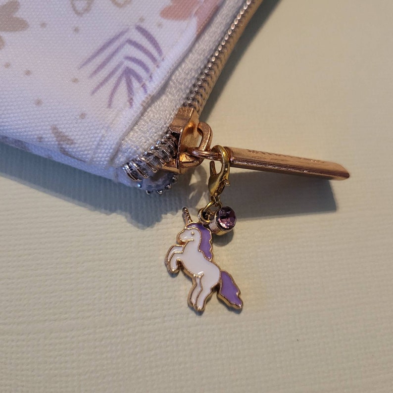 May include: A white zippered pouch with a floral pattern and a gold zipper pull. The zipper pull has a small unicorn charm with a purple mane and a pink gem.