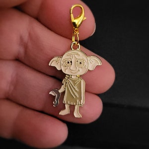 May include: Gold-colored charm featuring a cartoon Dobby the house-elf from the Harry Potter series. Dobby is wearing a brown robe and has a sad expression on his face.