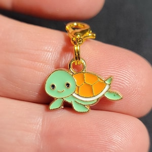 May include: A gold-coloured charm featuring a green and orange turtle design. The turtle has a smiling face and a white outline. The charm is attached to a gold-coloured lobster clasp.