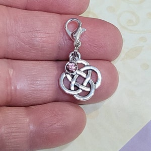May include: Silver charm featuring a pink crystal at its centre and a Celtic knot design. The charm has a lobster claw clasp.