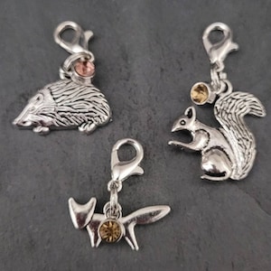 May include: Three silver charm clips with animal designs. The charms are a hedgehog, a squirrel, and a fox. Each charm has a small crystal in the center.