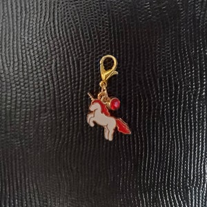 May include: A gold-coloured charm featuring a white unicorn with a red mane and a red gem. The charm is attached to a gold-coloured clip.