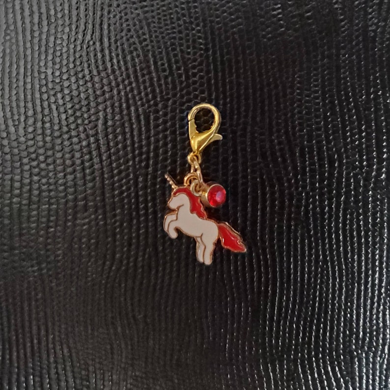 May include: A gold-coloured charm featuring a white unicorn with a red mane and a red gem. The charm is attached to a gold-coloured clip.