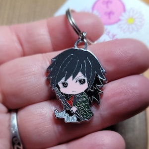 May include: A silver keychain featuring a cartoon character with black hair, a green and red outfit, and a sword. The character has a light pink face and dark eyes.