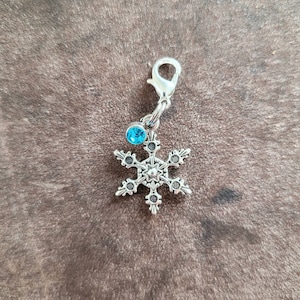 May include: Silver snowflake charm with a blue crystal accent. The charm has a lobster claw clasp for attaching to a bag, keychain, or other items.