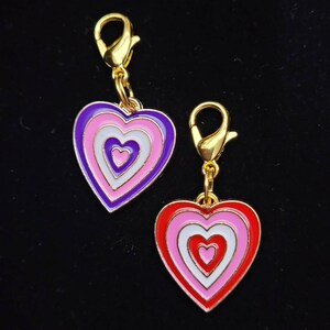 May include: Two gold heart-shaped charms with a clip at the top. The hearts are layered with concentric circles of pink, white, and purple or red.