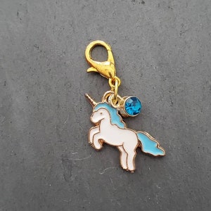 May include: Gold-tone charm featuring a white unicorn with a blue mane and tail, accented with a blue rhinestone. The charm has a lobster clasp.
