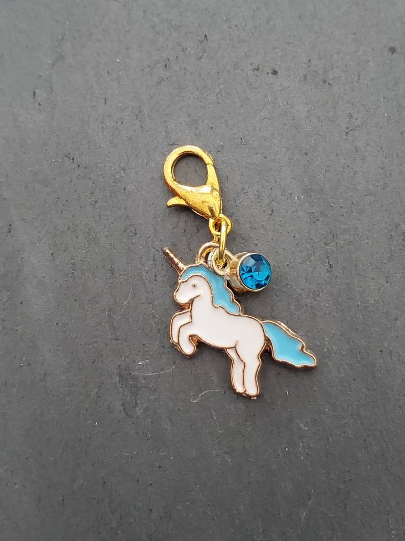 May include: Gold-tone charm featuring a white unicorn with a blue mane and tail, accented with a blue rhinestone. The charm has a lobster clasp.