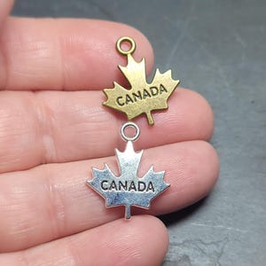 May include: Two metal maple leaf charms with the word "CANADA" inscribed. One charm is gold-toned, the other is silver-toned. Both charms have a small loop at the top for attaching to jewelry or other crafts.