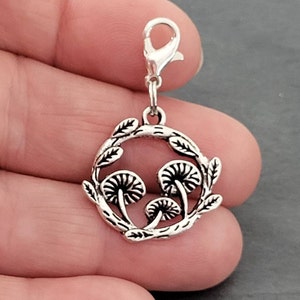May include: A silver charm depicting three mushrooms encircled by a wreath of leaves. The charm is attached to a lobster clasp. The design is detailed and has a vintage aesthetic, suitable for a bracelet or necklace.