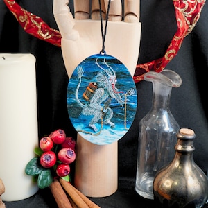 Op de afbeelding: Een ronde blauwe en witte kerstbal met een wit, harig, gehoornd wezen met een lange tong en een emmer op zijn rug. Het wezen staat op een besneeuwde ondergrond met een ketting die aan zijn pols is bevestigd.