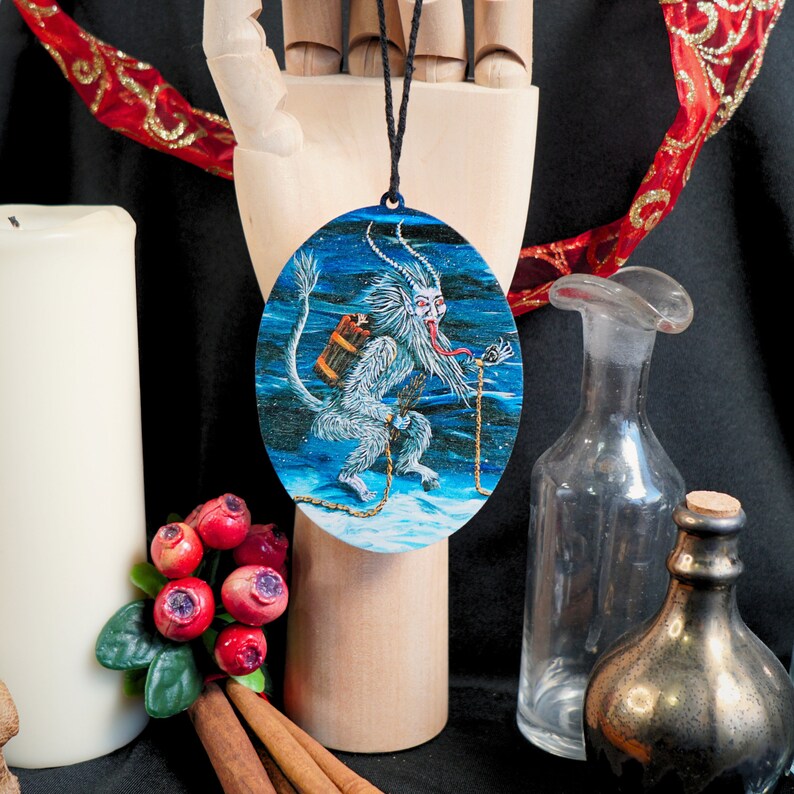 Op de afbeelding: Een ronde blauwe en witte kerstbal met een wit, harig, gehoornd wezen met een lange tong en een emmer op zijn rug. Het wezen staat op een besneeuwde ondergrond met een ketting die aan zijn pols is bevestigd.