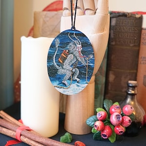 Op de afbeelding: Een ronde houten ornament met een blauwe en witte illustratie van een wit, harig wezen met rode hoorns en een lange rode tong die een rood pakket draagt. Het wezen loopt op een besneeuwd oppervlak met een touw aan zijn been.
