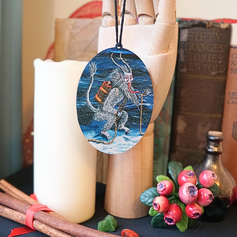Op de afbeelding: Een ronde houten ornament met een blauwe en witte illustratie van een wit, harig wezen met rode hoorns en een lange rode tong die een rood pakket draagt. Het wezen loopt op een besneeuwd oppervlak met een touw aan zijn been.
