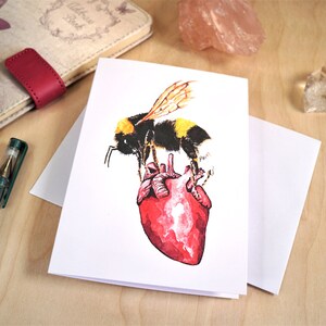 May include: A greeting card featuring a watercolour illustration of a bumblebee standing on a red heart.