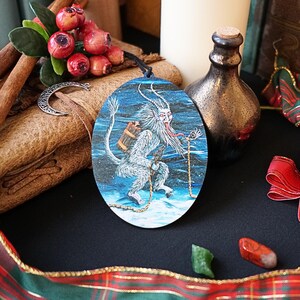 Op de afbeelding: Een ronde houten ornament met een blauwe en witte illustratie van een wit, harig wezen met een lange, rode tong en een ketting. Het wezen houdt een klein, bruin vat vast en loopt op een besneeuwde ondergrond. De ornament is versierd met een gouden ketting en een ster.