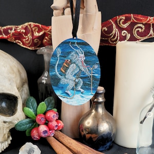 Op de afbeelding: Een ronde, blauw-witte kerstbal met een wit, harig wezen met een lange, rode tong en een vat op zijn rug. Het wezen staat op een besneeuwde ondergrond met een ketting.