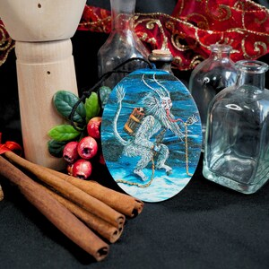 Op de afbeelding: Een ronde, blauw-witte kerstbal met een wit, harig wezen met rode ogen en een lange, rode tong. Het wezen houdt een klein, bruin tasje en een ketting vast. De kerstbal hangt aan een touwtje.