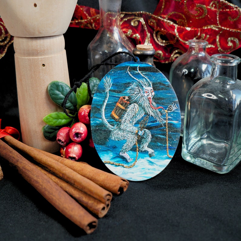 Op de afbeelding: Een ronde, blauw-witte kerstbal met een wit, harig wezen met rode ogen en een lange, rode tong. Het wezen houdt een klein, bruin tasje en een ketting vast. De kerstbal hangt aan een touwtje.