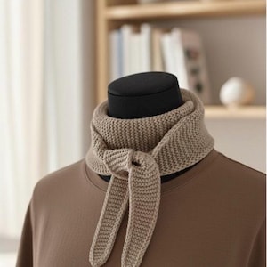 Könnte beinhalten: Ein beigefarbener Strickschal wird auf einer Schaufensterpuppe präsentiert. Der Schal ist vorne geknotet, mit zwei langen, rechteckigen Enden, die herunterhängen. Der Schal ist aus einem dicken, strukturierten Garn gefertigt. Die Schaufensterpuppe trägt ein braunes T-Shirt.