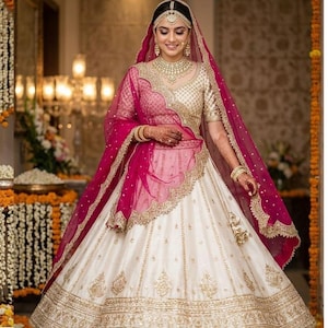 May include: A bride in a traditional Indian wedding outfit. The ensemble includes a cream-colored skirt with gold embroidery, a gold top, and a pink veil. The bride is adorned with gold jewelry, including a necklace, earrings, and headpiece. The background is softly lit.