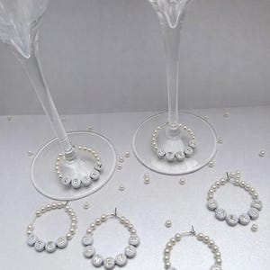 May include: Champagne glasses with pearl and lettered wine charms. The charms spell out names and words like "Bride" and "Sister". Scattered pearls add to the elegant, celebratory theme. Ideal for a wedding or bridal shower.