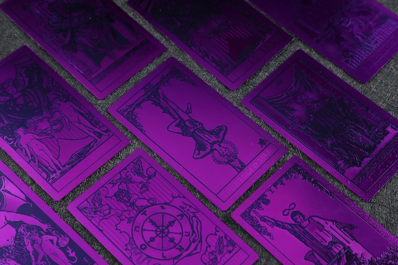 Pu&ograve; includere: Un set di carte dei tarocchi con lamina metallica viola e inchiostro blu. Le carte presentano varie immagini dai Tarocchi Maggiori, tra cui L'Impiccato, La Ruota della Fortuna e Il Carro.
