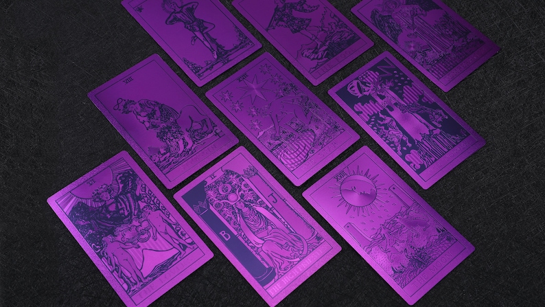 Pu&ograve; includere: Un set di nove carte dei tarocchi con sfondi metallici viola e illustrazioni in bianco e nero. Le carte sono disposte a ventaglio su una superficie grigio scuro.