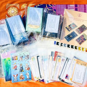 May include: A collection of tarot card decks and sticker sheets individually packaged in clear plastic sleeves. The items include blue and green boxes, a tan folder labeled The Wheel of Fortune, and a sheet of colorful cartoon rabbit stickers.