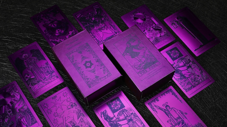 Pu&ograve; includere: Un set di carte dei tarocchi con accenti in lamina metallica viola. Le carte sono disposte a ventaglio, con la scatola al centro. La scatola &egrave; anche viola metallizzato con un disegno di una stella e un cerchio.