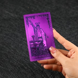 Pu&ograve; includere: Una carta dei tarocchi viola con finitura metallica. La carta raffigura una figura che tiene una bacchetta con un simbolo di infinito sopra la sua testa. La carta &egrave; etichettata come "Il Mago".