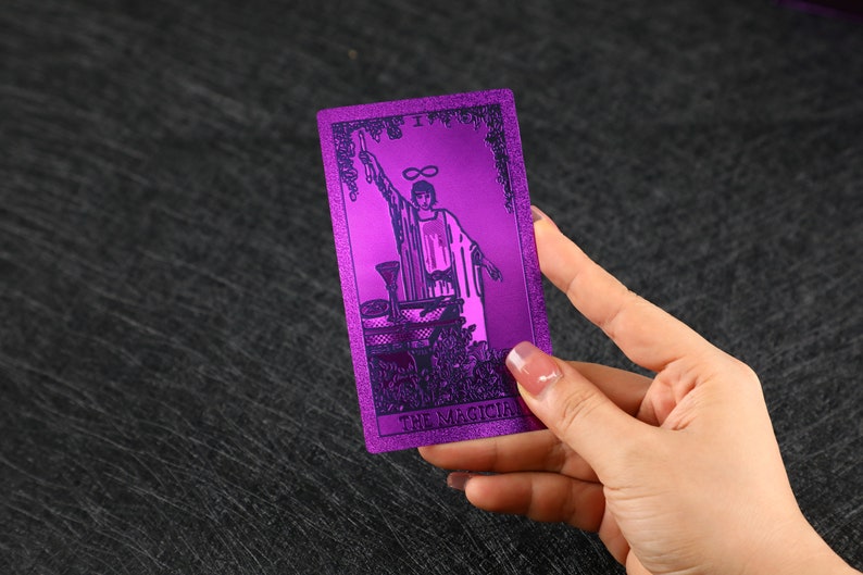 Pu&ograve; includere: Una carta dei tarocchi viola con finitura metallica. La carta raffigura una figura che tiene una bacchetta con un simbolo di infinito sopra la sua testa. La carta &egrave; etichettata come "Il Mago".