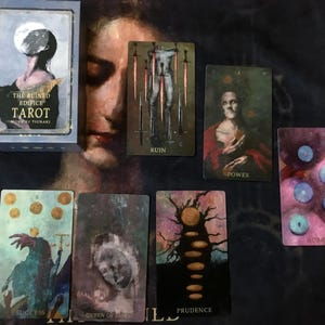 May include: A tarot card deck titled "The Ruined Edifice Tarot" with several cards displayed. The cards feature illustrations with words like "Ruin," "Power," "Success," and "Prudence." The deck box is light blue with a moon design.