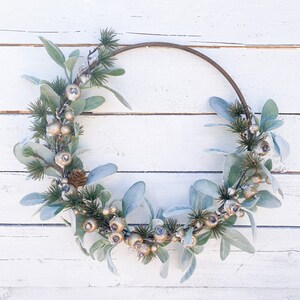 May include: A circular wreath with a brown metal ring base, adorned with silver-colored berries, small pine cones, and soft, gray-green leaves. The wreath is set against a white, weathered wooden background.