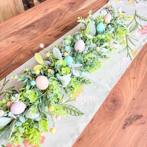 Könnte beinhalten: Dekorativer Ostern-Tischläufer mit gestickten Blumenmustern in Rosa, Gelb und Grün. Der Läufer ist mit einer Girlande aus grünem Laub und pastellfarbenen Ostereiern geschmückt, was eine festliche Frühlingsdekoration ergibt.