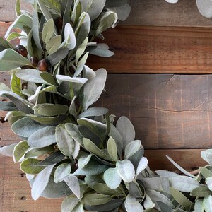 May include: A green and gray artificial lamb's ear wreath with brown berries. The wreath is on a rustic wooden background.
