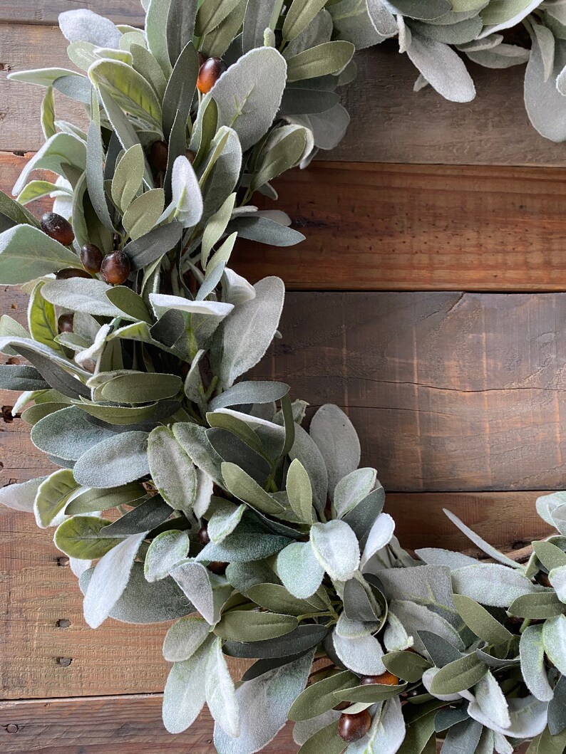 May include: A green and gray artificial lamb's ear wreath with brown berries. The wreath is on a rustic wooden background.