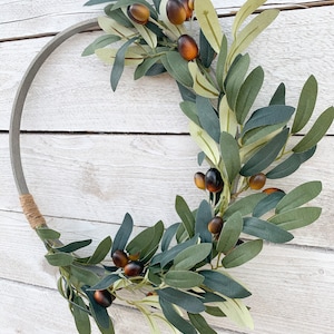 May include: Olive branch wreath with brown olives and green leaves on a white wooden background.
