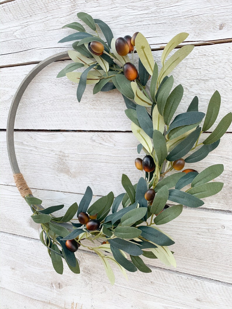 May include: Olive branch wreath with brown olives and green leaves on a white wooden background.