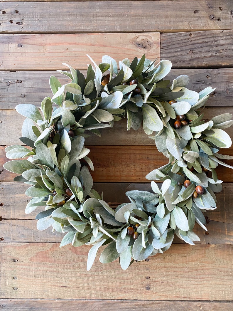 May include: A green and brown wreath made of faux lambs ear leaves and brown berries. The wreath is hanging on a wooden wall.