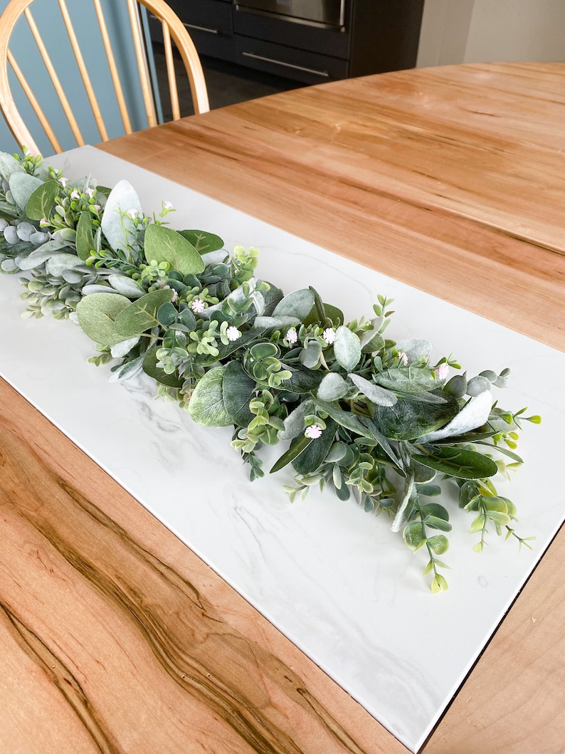 Puede incluir: Un camino de mesa de m&aacute;rmol blanco con una guirnalda verde y blanca de vegetaci&oacute;n artificial y peque&ntilde;as flores blancas.