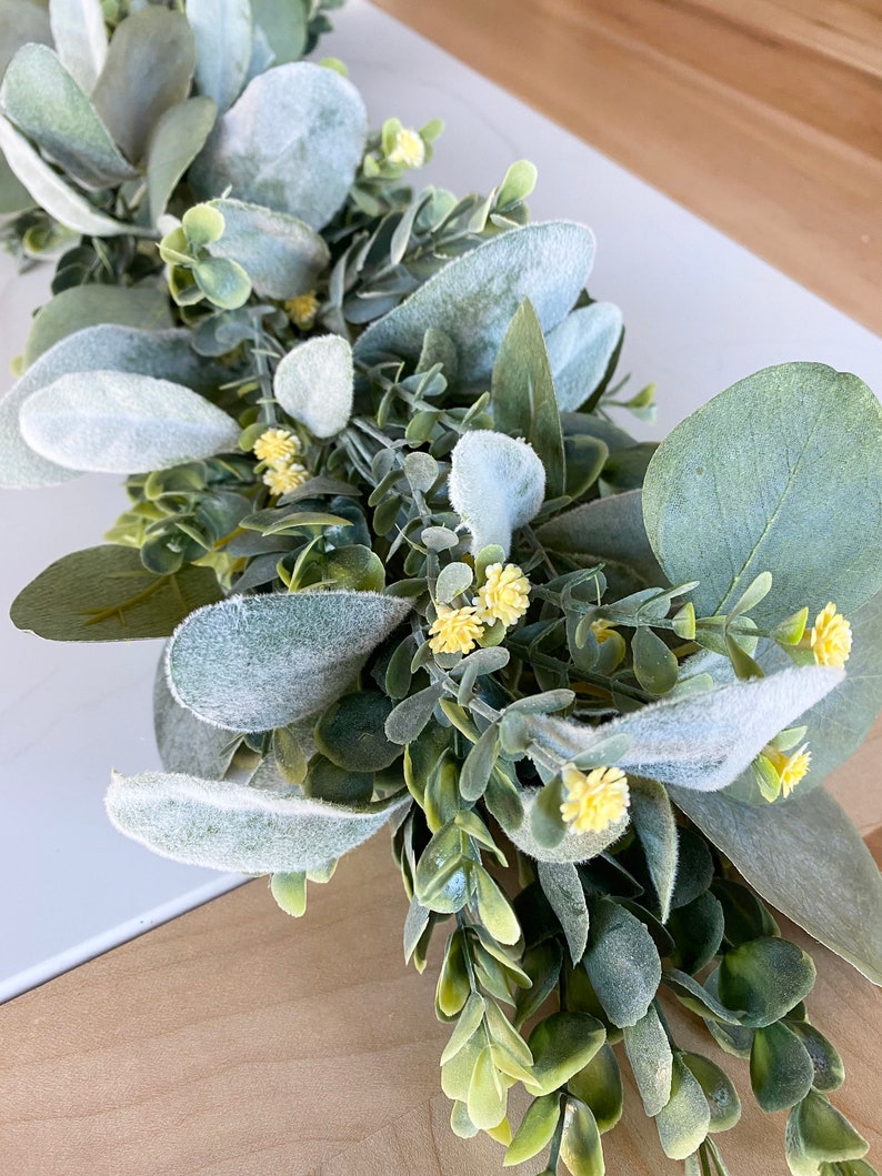 Puede incluir: Una guirnalda artificial de eucalipto verde y gris con peque&ntilde;as flores amarillas. La guirnalda est&aacute; hecha de hojas y flores falsas y es perfecta para agregar un toque de vegetaci&oacute;n a la decoraci&oacute;n de tu hogar.