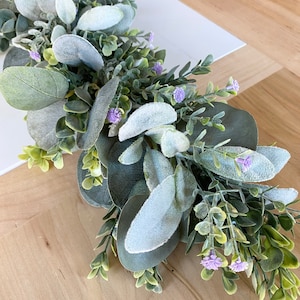 Puede incluir: Una guirnalda de eucalipto verde y gris con peque&ntilde;as flores moradas. La guirnalda es perfecta para a&ntilde;adir un toque de vegetaci&oacute;n a la decoraci&oacute;n de tu hogar.