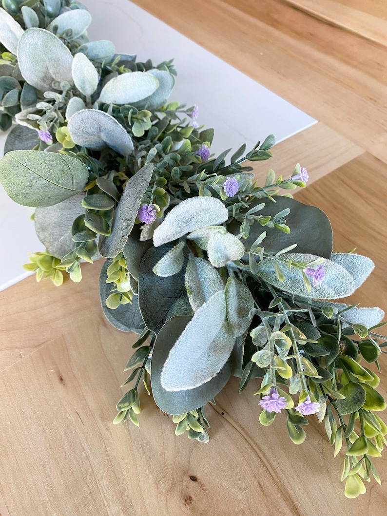 Puede incluir: Una guirnalda de eucalipto verde y gris con peque&ntilde;as flores moradas. La guirnalda es perfecta para a&ntilde;adir un toque de vegetaci&oacute;n a la decoraci&oacute;n de tu hogar.