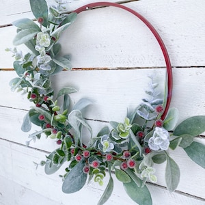May include: A red metal wreath with frosted eucalyptus leaves and red berries. The wreath is hanging on a white wooden wall.