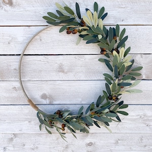 May include: A green olive branch wreath with brown olives, arranged in a circle on a white wooden background.